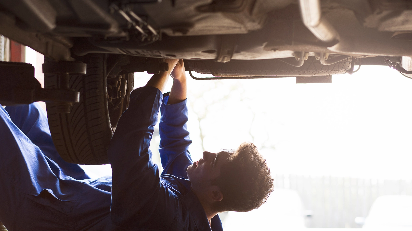 Junger Mann liegt im Blaumann unter repariert etwas am Unterboden.